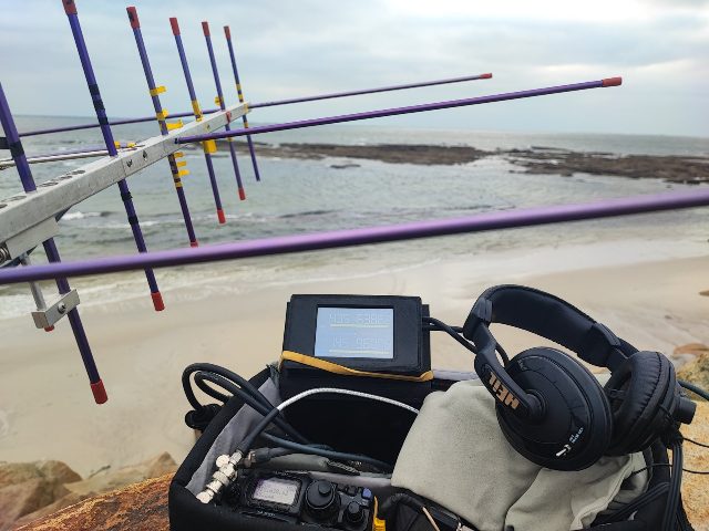 The bag open on the beach: the FT-817, a small screen showing the 435 and 145 MHz frequencies, the Heil headset, and the crossed Yagi — December 2025